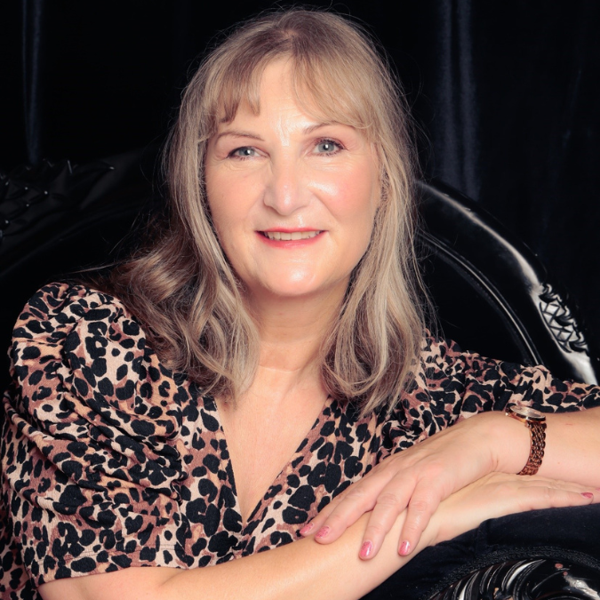 A portrait of a woman with shoulder-length blonde hair, smiling gently. She is wearing a leopard-print blouse and has her arms crossed in front of her, resting on a black, ornate chair. The background is a dark fabric, creating a soft contrast with her light skin and patterned blouse.